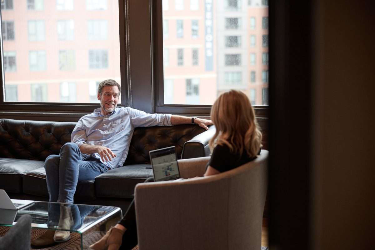 A bearded man in a button down shirt talks with a blonde haired woman on a leather sofa in the CTP office.